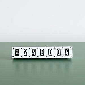 Może przedstawiać: Biało-czarny wyświetlacz z klapkami z numerami 8248004. Prostokątny wyświetlacz stoi na zielonej powierzchni. Tło to gładka biała ściana.