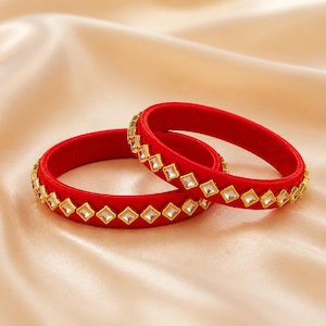 Puede incluir: Dos brazaletes circulares envueltos en hilo rojo vibrante, cada uno con una fila continua de pequeñas piedras claras de forma cuadrada engastadas en marcos de metal dorado que rodean la superficie exterior de las piezas.