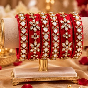 May include: A set of six red bangles featuring a vibrant fabric texture, adorned with clear, diamond-shaped mirror accents and small, circular crystal embellishments arranged in floral patterns along the outer surface of each circular bracelet.