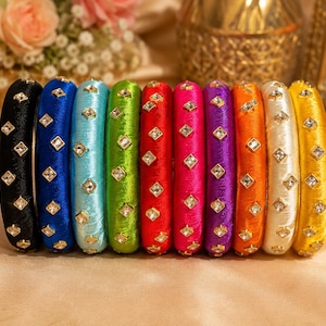 May include: A set of ten cylindrical bangles arranged in a row, each wrapped in vibrant silk thread in colors including black, blue, turquoise, green, red, pink, purple, orange, white, and yellow, featuring embedded square-cut clear crystals.