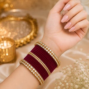 Peut inclure: Un ensemble de bracelets rigides en velours bordeaux profond empilés avec des bracelets en métal doré ornés de pierres claires carrées. L'ensemble comprend deux bracelets dorés extérieurs à pierres carrées, un fin bracelet perlé central.