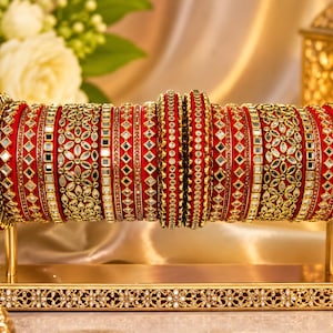 May include: A set of red bangles arranged on a gold-toned display stand. The bangles feature intricate gold-coloured metalwork, including floral patterns, geometric shapes, and embedded reflective accents that create a shimmering, textured appearance.