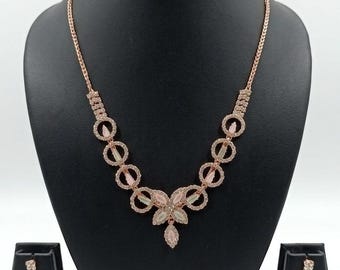 Op de afbeelding: Een roségoudkleurige ketting- en oorbellenset met een centraal bloemmotief van lichtroze en heldere kristallen. De ketting is verbonden met cirkelvormige schakels bezet met kleine steentjes, terwijl de bijpassende oorbellen een cirkelvormig ontwerp hebben met een enkele bungelende kristallen druppel.