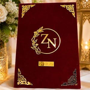 Könnte beinhalten: Ein rechteckiges Schild aus tiefrotem Samt mit goldfarbenen metallischen Akzenten. In der Mitte befinden sich die Buchstaben ZN in einem kreisförmigen Blumenkranz, ergänzt durch dekorative Eckverzierungen und ein Schild mit dem Datum 21.01.25.