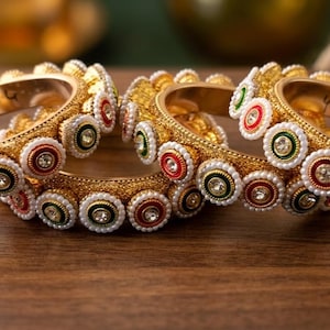 Può includere: Tre bracciali rigidi color oro con una fascia in metallo testurizzato decorata da un motivo ripetitivo di cerchi. Ogni motivo presenta una pietra centrale trasparente circondata da anelli in smalto bianco, rosso e verde e piccole perline bianche.