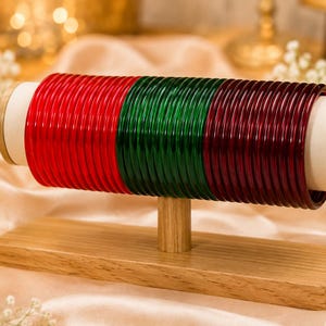 Op de afbeelding: Een set dunne, ronde armbanden gerangschikt op een horizontale houten displaystandaard. De armbanden zijn georganiseerd in drie verschillende kleurgroepen: helderrood, smaragdgroen en donkerrood, allemaal met een gladde, glanzende en doorschijnende afwerking.