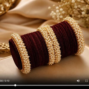 Op de afbeelding: Een set donkerrode fluwelen armbanden gerangschikt op een cilindrische houder. De armbanden zijn voorzien van ingewikkelde randen van kleine, crèmekleurige kunstparels, met twee bredere parelbanden die de fluwelen secties van elkaar scheiden.