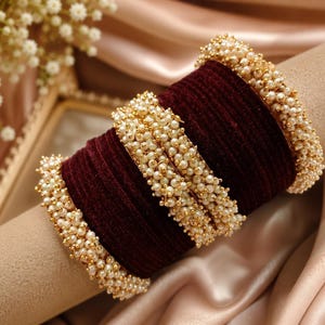 May include: A set of deep maroon velvet bangles arranged in a stack. The bangles are accented with rows of small, round, cream-coloured faux pearls set in gold-toned metal, featuring pearl-encrusted edges and two central pearl-adorned bands.