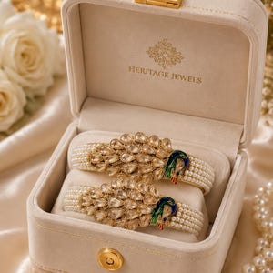 May include: A pair of gold-toned bangles featuring a peacock motif with clear teardrop-shaped stones, green and blue enamel detailing, and rows of small white pearls. The jewelry is presented in a beige velvet-lined box labeled Heritage Jewels.