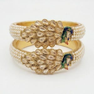 May include: Two gold-toned bangles featuring a central peacock motif adorned with clear, teardrop-shaped stones and enamel detailing in blue, green, and red. The bands are bordered by rows of small, white, spherical faux pearls.
