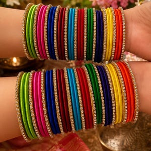 May include: A set of multi-colored silk thread bangles featuring vibrant shades of lime green, hot pink, royal blue, deep red, turquoise, emerald green, yellow, and orange, each accented with a thin, sparkling gold-tone rhinestone trim.