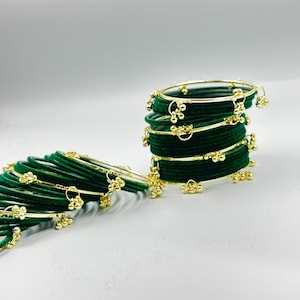 May include: A collection of dark green velvet-textured bangles arranged in a stack and a horizontal row. Each bangle features small, dangling gold-toned metal charms attached to the outer edge, creating a decorative and uniform appearance.