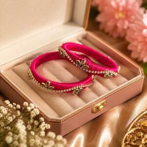 May include: Two circular bangles wrapped in vibrant fuchsia thread, each featuring a delicate row of clear rhinestones and small, leaf-shaped metallic accents arranged in a repeating pattern along the outer surface of the jewellery.