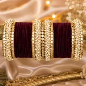Op de afbeelding: Een set armbanden met diep bordeauxrode fluweelachtige banden afgewisseld met goudkleurige metalen banden ingelegd met kleine witte parels en vierkante heldere stenen, symmetrisch gerangschikt op een horizontale displaystang.