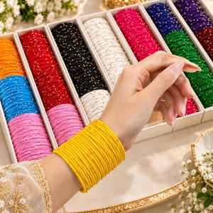 May include: A collection of thin, round bangles in various solid colors including yellow, orange, red, black, white, pink, blue, green, and purple. Each bangle features a smooth finish with small, evenly spaced gold-colored metallic accents.
