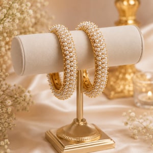 May include: Two circular bangles featuring a dense, multi-row arrangement of small, round, white faux pearls set against a gold-toned metal base. The bangles are displayed on a cylindrical stand, showcasing the uniform texture and reflective finish.