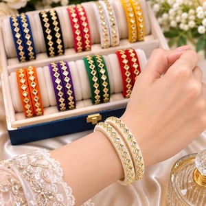 May include: A collection of colorful bangles featuring a central row of diamond-shaped mirrors set in gold-toned frames. The set includes pairs in blue, black, pink, white, yellow, orange, purple, green, and red, with cream-colored bangles worn.