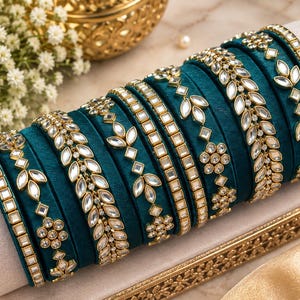 May include: A set of teal velvet bangles arranged on a cylindrical display. Each bangle features gold-toned metal accents with clear, reflective, leaf-shaped and circular stones arranged in intricate patterns along the surface of the fabric.