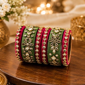 May include: A set of six bangles featuring alternating green and magenta fabric-wrapped bands. The green bangles are decorated with clear, leaf-shaped stones in a floral pattern, while the magenta bangles feature a single row of square stones.