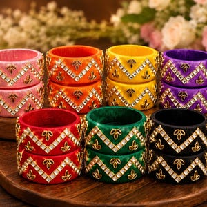 May include: A collection of eight cylindrical bangles in pink, orange, yellow, purple, red, green, and black. Each bangle features a zigzag pattern of embedded clear stones and small gold-toned leaf-shaped accents along the outer surface.
