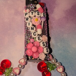 May include: A decorated lighter with the text "PTSD PRINCESS" in white letter beads. The lighter is embellished with pink flowers, rhinestones, and pill-shaped charms. A beaded cherry and heart charm hangs from the base.