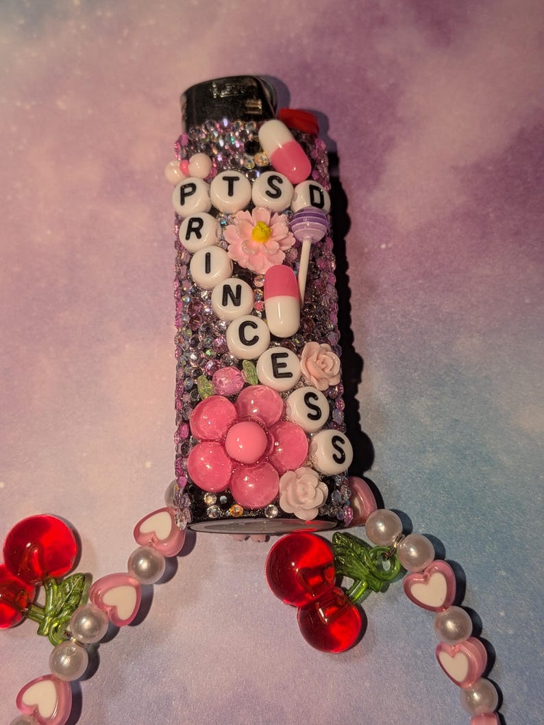 May include: A decorated lighter with the text "PTSD PRINCESS" in white letter beads. The lighter is embellished with pink flowers, rhinestones, and pill-shaped charms. A beaded cherry and heart charm hangs from the base.