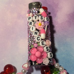 May include: A decorative lighter covered in pink rhinestones and adorned with pink flowers and pill-shaped beads. The word "PTSD" is spelled out in white letter beads. A beaded charm with cherries and hearts hangs from the bottom.
