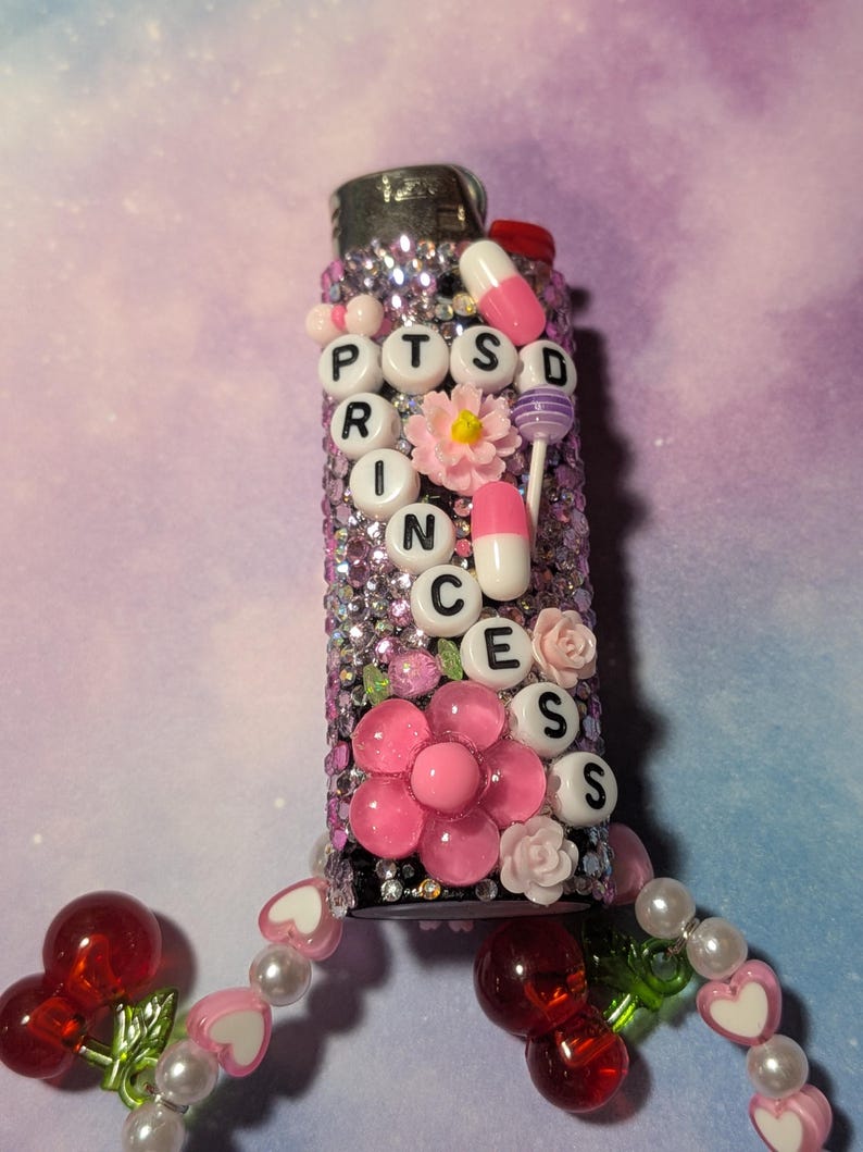 May include: A decorative lighter covered in pink rhinestones and adorned with pink flowers and pill-shaped beads. The word "PTSD" is spelled out in white letter beads. A beaded charm with cherries and hearts hangs from the bottom.