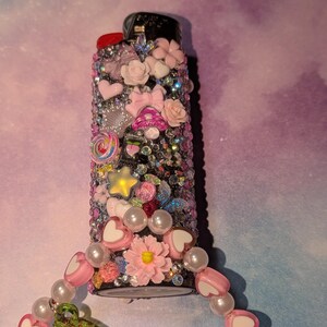 May include: A decorative lighter covered in rhinestones and embellished with pink hearts, bows, flowers, and a star. A beaded charm with cherries and heart-shaped beads is attached to the lighter.