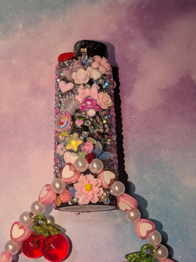 May include: A decorative lighter covered in rhinestones and embellished with pink hearts, bows, flowers, and a star. A beaded charm with cherries and heart-shaped beads is attached to the lighter.