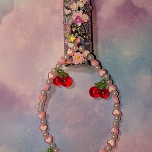 May include: A decorated lighter covered in rhinestones, flowers, and charms. A beaded cherry charm bracelet is attached. The lighter has a red button and a silver metal top.