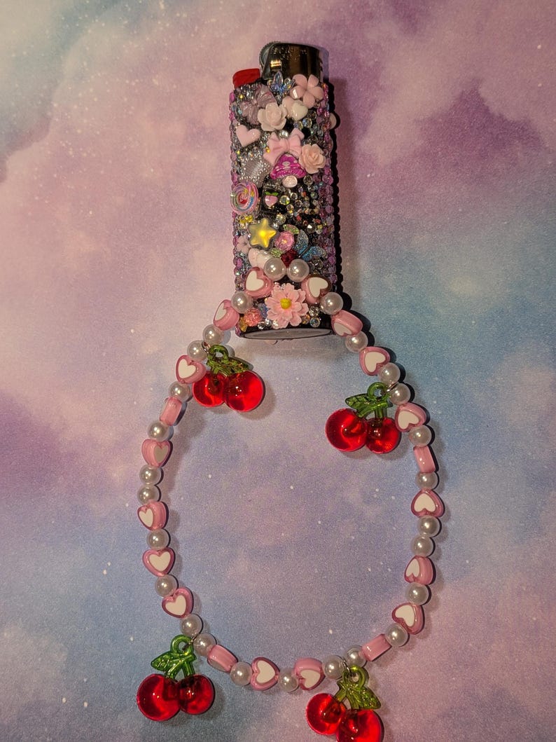 May include: A decorated lighter covered in rhinestones, flowers, and charms. A beaded cherry charm bracelet is attached. The lighter has a red button and a silver metal top.