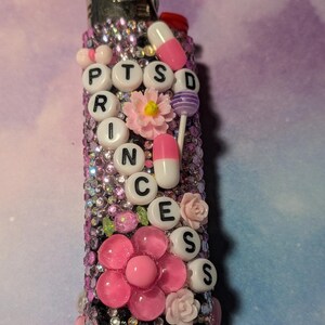 May include: A decorated lighter featuring the text "PTSD PRINCESS" in white beads. Adorned with pink flowers, faux pills, and rhinestones. A distinctive, personalised accessory. The lighter is approximately 8 cm in length.
