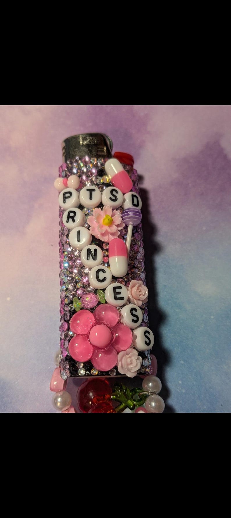 May include: A decorated lighter featuring the text "PTSD PRINCESS" in white beads. Adorned with pink flowers, faux pills, and rhinestones. A distinctive, personalised accessory. The lighter is approximately 8 cm in length.