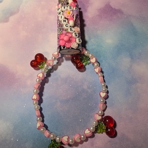 May include: A decorative lighter with the word "PRINCESS" spelled out in letter beads. The lighter is embellished with pink flowers, glitter, and beads. A beaded bracelet with heart and pearl beads and red cherry charms is attached.