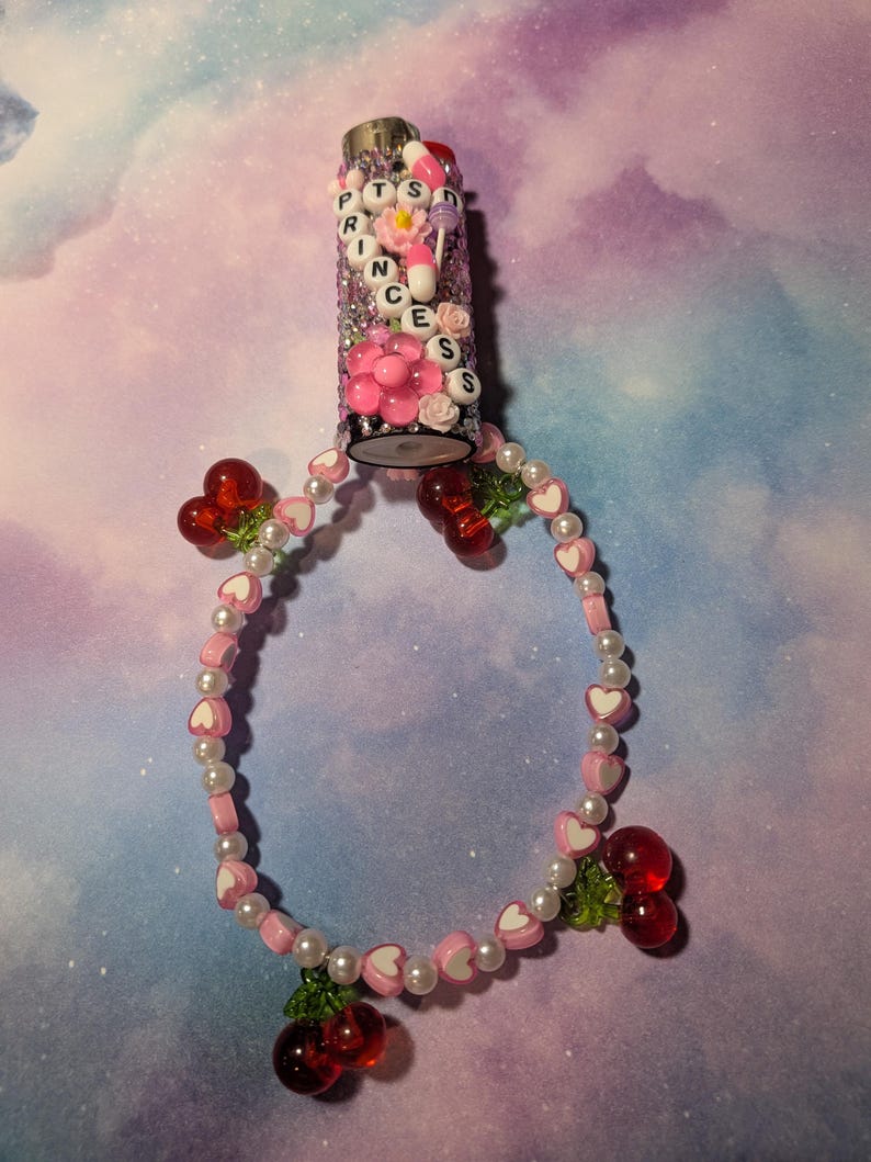 May include: A decorative lighter with the word "PRINCESS" spelled out in letter beads. The lighter is embellished with pink flowers, glitter, and beads. A beaded bracelet with heart and pearl beads and red cherry charms is attached.