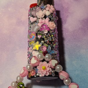 May include: A decorative lighter covered in rhinestones and adorned with pink bows, hearts, and flowers. A charm with pink heart beads, pearls, and red cherry charms is attached. The lighter is a unique, embellished accessory.