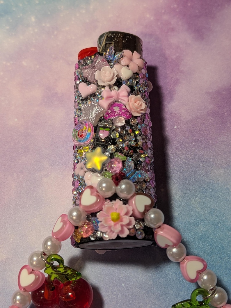 May include: A decorative lighter covered in rhinestones and adorned with pink bows, hearts, and flowers. A charm with pink heart beads, pearls, and red cherry charms is attached. The lighter is a unique, embellished accessory.