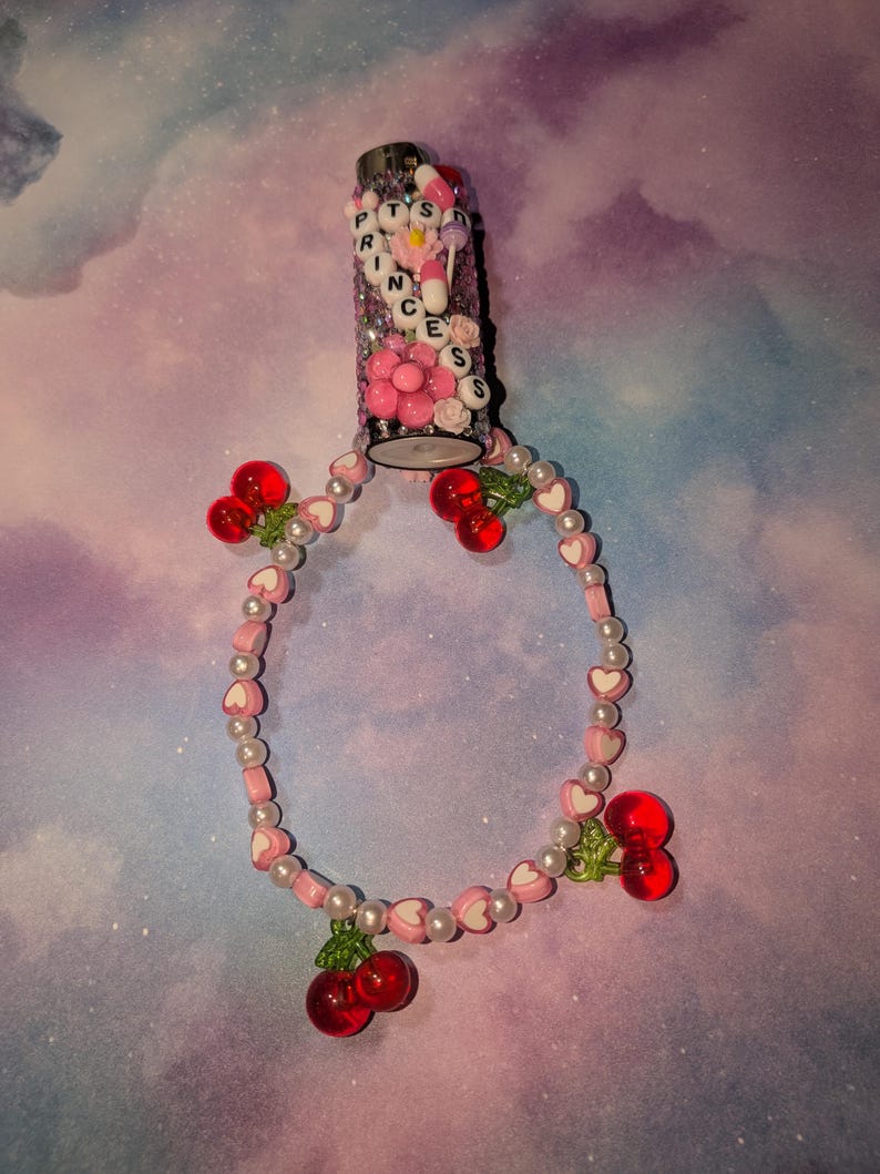 May include: A glitter-covered lighter with the word "PRINCESS" spelled out in letter beads. A pink and white beaded bracelet with heart-shaped beads and red cherry charms is attached to the lighter.