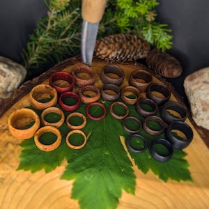May include: A selection of handcrafted wooden rings in various sizes and wood grains, including light, medium, and dark tones. The rings are displayed on a green leaf and a wooden surface, with a carving tool and natural elements in the background.