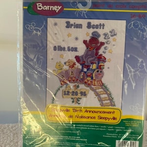 May include: A Barney cross-stitch kit featuring a colorful design with Barney the dinosaur, stars, and the text "Brian Scott" and "12-26-96". The kit includes the text "Sleepyville Birth Announcement".