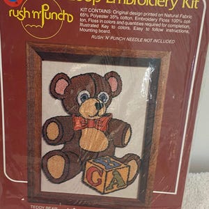 May include: A "Boye" brand Loop Embroidery Kit featuring a teddy bear design. The kit includes a framed image of a brown teddy bear with a red bow tie and a block with the letters "GCA". The kit fits a standard 5"x7" frame.