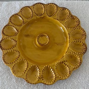 May include: A mustard-yellow ceramic serving platter with a scalloped edge and a central circular indentation. The platter features 16 decorative, fan-shaped indentations around the rim, and a slightly darker, aged appearance around the edges.