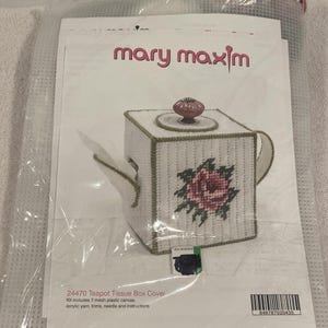 May include: A Mary Maxim teapot tissue box cover kit. The kit includes a white, square tissue box cover with a pink rose design and a brown knob on top. The text "mary maxim" is visible.