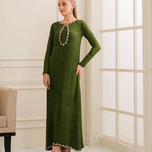 Könnte beinhalten: Olivgrünes Langarmkleid mit Schlüsselloch-Ausschnitt und Verzierungen. Das Kleid hat kleine, verstreute Verzierungen und eine dekorative Bordüre am Ausschnitt und am Saum. Das Kleid ist bodenlang.