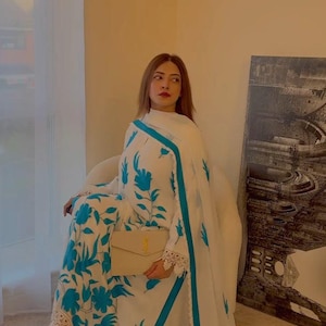 Könnte beinhalten: Ein weißes Ensemble mit blauen Blumenmustern, einem passenden Schal und einer beigen Clutch. Das Outfit hat eine Spitzenborte und blaue Akzente. Die Person sitzt auf einem Stuhl.