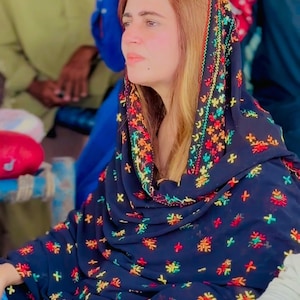 Puede incluir: Una mujer con un pañuelo azul oscuro con bordados florales coloridos. El pañuelo está drapeado sobre la cabeza y los hombros, con un diseño bordado con flores rojas, amarillas y verdes. La mujer lleva un reloj.