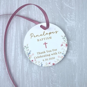 May include: White round favor tag with a floral design and gold lettering. The tag says "Penelope's Baptism" with a pink cross and "Thank You For Celebrating with Us 4.30.2030".