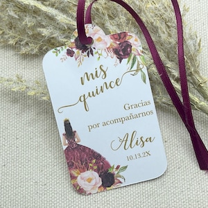 May include: A white card with gold lettering and floral designs. The text reads "mis quince" and "Gracias por acompañarnos Alisa 10.13.2X".