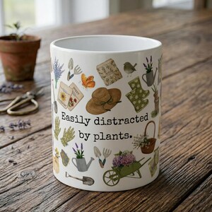Puede incluir: Taza de cerámica blanca con un diseño de jardín. La taza está decorada con ilustraciones de herramientas de jardinería, plantas y el texto "Easily distracted by plants". Una pequeña maceta de terracota con una planta y tijeras de podar en el fondo.